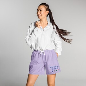 May include: A white long-sleeve quarter-zip hooded jacket with a drawstring hem paired with light purple Fog athletic shorts. The shorts by Alter Standard feature the blue text MONTRÉAL CANADA above four small colorful icons including a fleur-de-lis and a rose.
