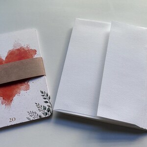 May include: A stack of white rectangular cards featuring a red watercolour heart and black botanical illustrations at the bottom, secured by a brown paper band with the number 20 printed on the card, alongside a stack of plain white envelopes.