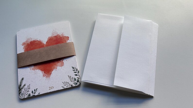 May include: A stack of white rectangular cards featuring a red watercolour heart and black botanical illustrations at the bottom, secured by a brown paper band with the number 20 printed on the card, alongside a stack of plain white envelopes.