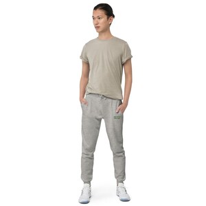 May include: A beige short-sleeved crew neck t-shirt and heather grey jogger sweatpants with a drawstring waist and ribbed cuffs. The sweatpants feature small green text on the upper left thigh area. The outfit is paired with white athletic trainers.