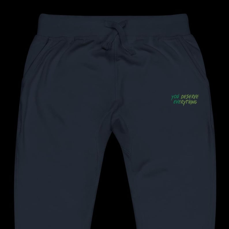 May include: Dark navy blue sweatpants featuring an elastic waistband with a matching drawstring. The right side displays the text "YOU DESERVE EVERYTHING" printed in a gradient of green and yellow letters in a casual, handwritten font style.
