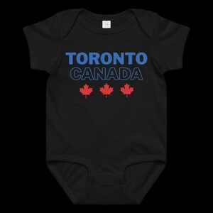 以下が含まれることがあります： 黒の半袖ベビー用ボディスーツ。青い文字で「TORONTO」、青い輪郭のフォントで「CANADA」と書かれ、その下に赤いカエデの葉のアイコンが3つ横一列に並んでいます。この衣服は丸首のデザインです。