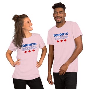 Op de afbeelding: Een lichtroze t-shirt met korte mouwen voorzien van de tekst TORONTO in het blauw, CANADA in een blauwe omlijning en drie rode esdoornbladpictogrammen op de borst. Het kledingstuk heeft een ronde hals en een eenvoudige pasvorm.