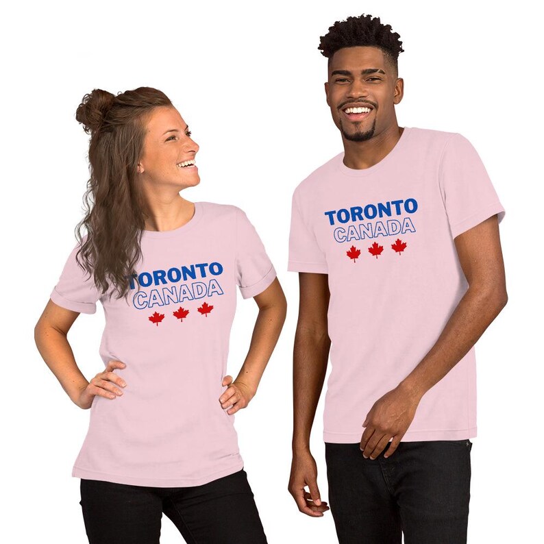 Op de afbeelding: Een lichtroze t-shirt met korte mouwen voorzien van de tekst TORONTO in het blauw, CANADA in een blauwe omlijning en drie rode esdoornbladpictogrammen op de borst. Het kledingstuk heeft een ronde hals en een eenvoudige pasvorm.