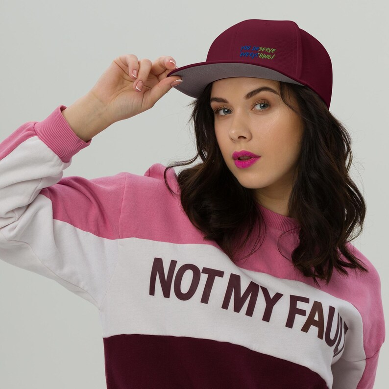 May include: A maroon baseball cap with a grey underside of the brim and the text "YOU DESERVE EVERYTHING!" embroidered in blue and green. The cap is paired with a colour-blocked sweatshirt featuring pink, white, and maroon panels with "NOT MY FAULT" text.