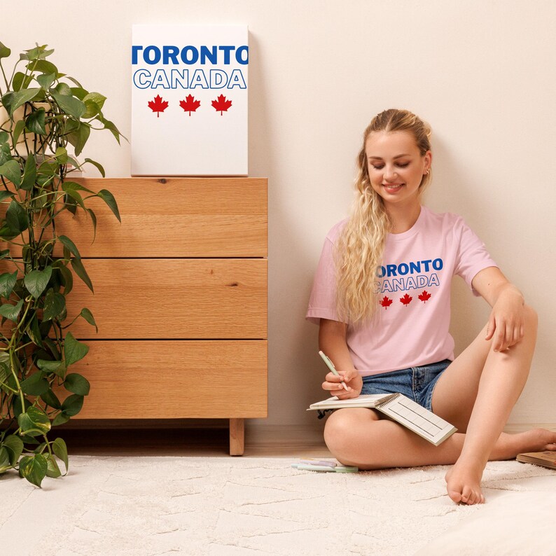 Op de afbeelding: Een lichtroze t-shirt met de blauwe tekst TORONTO en de wit omrande tekst CANADA, gevolgd door drie rode esdoornbladiconen. Hetzelfde grafische ontwerp staat op een wit rechthoekig bord op een houten ladekast.