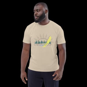 Pode incluir: Uma t-shirt bege de manga curta com uma impress&atilde;o gr&aacute;fica de uma paisagem florestal com montanhas, um design de raios de sol e o texto "COLLINGWOOD" com "44.5000&deg;N" impresso acima, acentuado por um tra&ccedil;o diagonal amarelo brilhante.