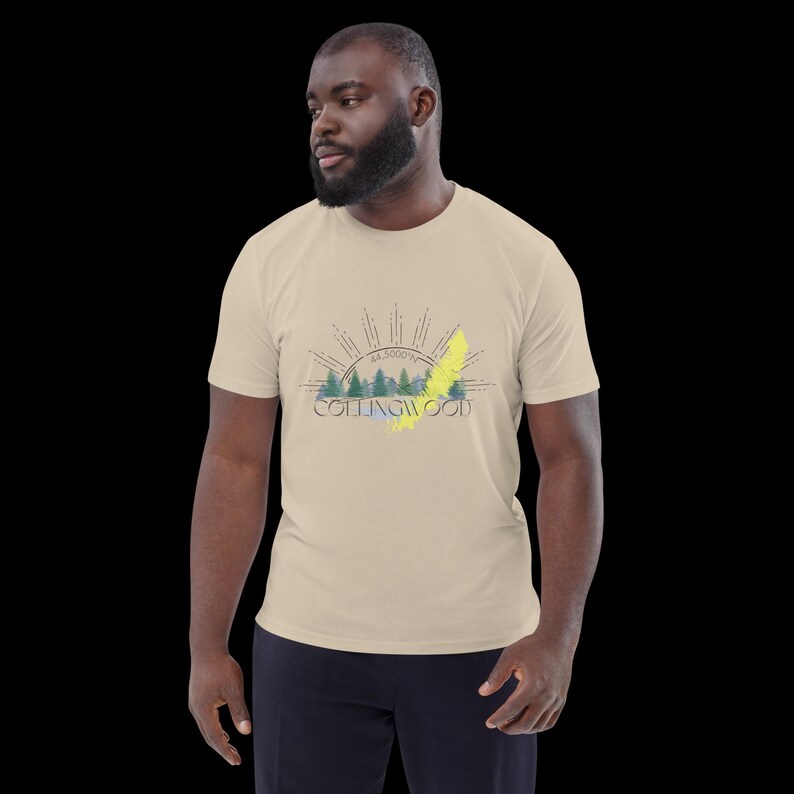 Pode incluir: Uma t-shirt bege de manga curta com uma impress&atilde;o gr&aacute;fica de uma paisagem florestal com montanhas, um design de raios de sol e o texto "COLLINGWOOD" com "44.5000&deg;N" impresso acima, acentuado por um tra&ccedil;o diagonal amarelo brilhante.