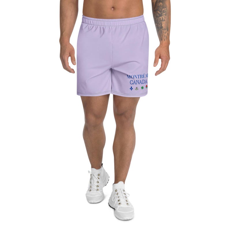 May include: Light purple athletic shorts featuring an elastic waistband and a drawstring. The left leg displays the blue text "MONTR&Eacute;AL CANADA" above four small, colorful floral and heraldic icons including a fleur-de-lis and a clover.