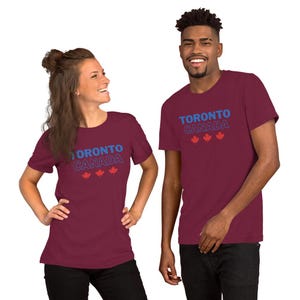 Op de afbeelding: Een bordeauxrood t-shirt met korte mouwen voorzien van de blauwe tekst TORONTO en de wit omlijnde tekst CANADA, vergezeld door drie kleine rode esdoornblad-iconen gecentreerd onder de tekst op de voorzijde van het kledingstuk.