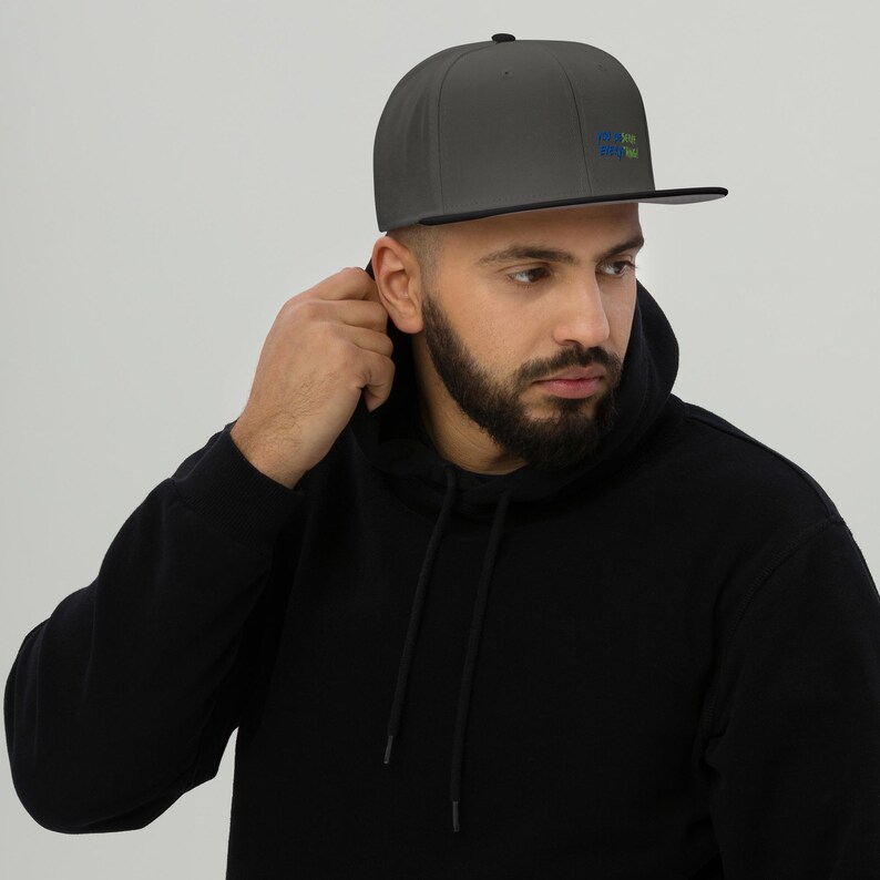 May include: A charcoal grey baseball cap with a flat brim and a small, multi-coloured graphic logo on the front panel. The cap features a structured crown and a solid dark grey fabric construction, providing a clean and simple aesthetic design.