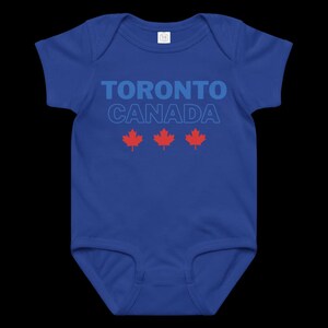 以下が含まれることがあります： ロイヤルブルーの半袖ベビー用ボディスーツ。太字の青いフォントで「TORONTO CANADA」とプリントされており、「CANADA」の文字は明るい青色で縁取られています。テキストの下には、3つの小さな赤いカエデの葉のアイコンが横一列に並んでいます。