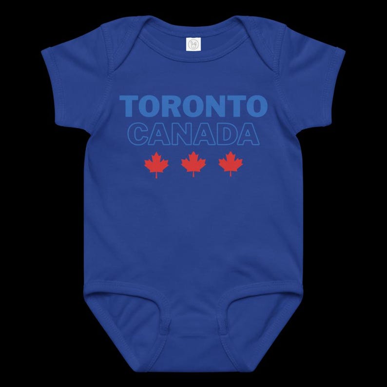 以下が含まれることがあります： ロイヤルブルーの半袖ベビー用ボディスーツ。太字の青いフォントで「TORONTO CANADA」とプリントされており、「CANADA」の文字は明るい青色で縁取られています。テキストの下には、3つの小さな赤いカエデの葉のアイコンが横一列に並んでいます。