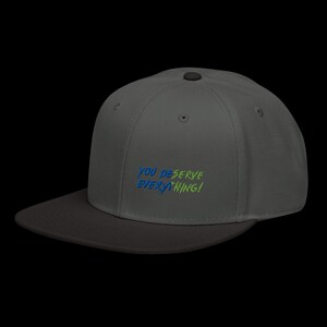 May include: A charcoal grey baseball cap with a flat brim and a top button. The front features embroidered text in blue and lime green that reads YOU DESERVE EVERYTHING! The cap has a structured crown with ventilation eyelets on the side panels.