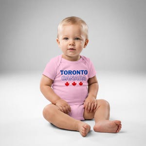 以下が含まれることがあります： 胸の中央に青い文字でTORONTO、白い縁取りの文字でCANADA、そして3つの小さな赤いカエデの葉のアイコンがプリントされた、ライトピンクの半袖ベビー用ボディスーツ。丸首で、股下にスナップボタンが付いています。