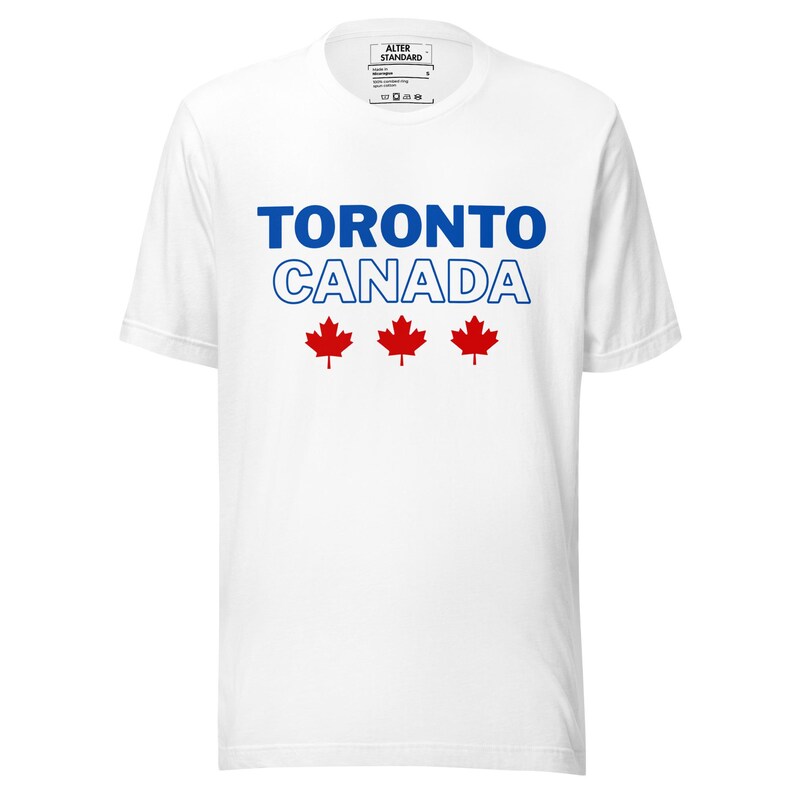 Op de afbeelding: Een wit t-shirt met korte mouwen voorzien van de blauwe tekst TORONTO en de blauw omlijnde tekst CANADA, gevolgd door drie rode esdoornblad-iconen in het midden op de borst. Het label in de nek toont de merknaam Alter Standard.