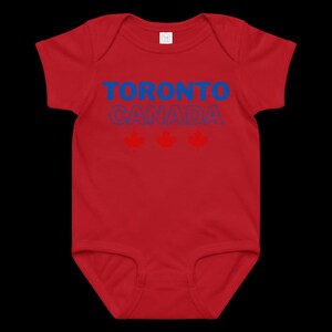 以下が含まれることがあります： 赤い半袖のベビー用ボディスーツ。前面には青い文字で「TORONTO」、青い輪郭の文字で「CANADA」とプリントされており、その下には3つの赤いカエデの葉のアイコンが水平に並んでいます。