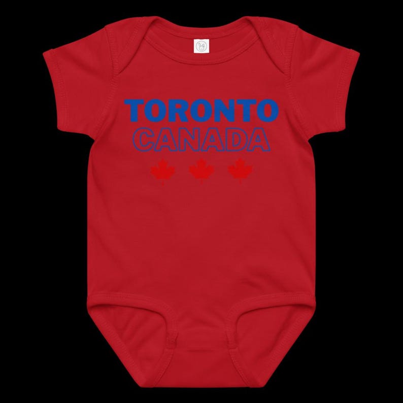 以下が含まれることがあります： 赤い半袖のベビー用ボディスーツ。前面には青い文字で「TORONTO」、青い輪郭の文字で「CANADA」とプリントされており、その下には3つの赤いカエデの葉のアイコンが水平に並んでいます。