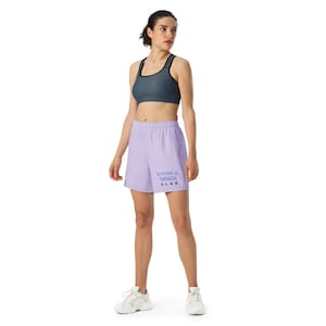 May include: A dark gray sports bra with black trim and light purple athletic shorts featuring the text MONTR&Eacute;AL CANADA in blue, accompanied by four small colorful floral icons printed on the lower left side of the shorts, paired with white sneakers.