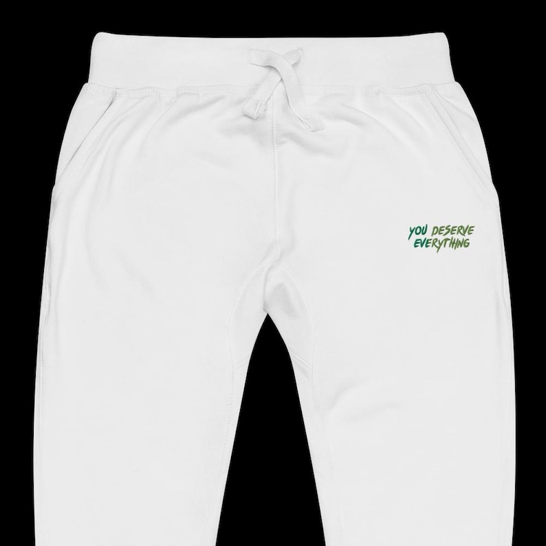 May include: White jogging bottoms featuring an elasticated waistband with a drawstring and the text "YOU DESERVE EVERYTHING" printed in a stylised green font on the upper right side. The garment has a clean, solid white finish throughout the fabric.