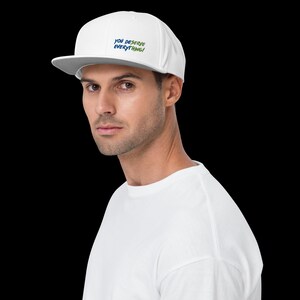 May include: A white baseball cap with a flat brim and the text YOU DESERVE EVERYTHING! embroidered in blue and green on the front panel. The cap features a structured crown and a matching white button on top, providing a clean and simple look.