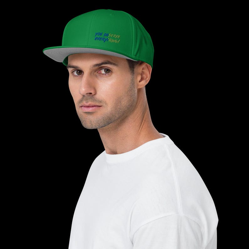 May include: A green baseball cap with a flat grey brim. The front of the cap features the embroidered text "YOU DESERVE EVERYTHING!" in blue and yellow lettering. The cap has a structured crown and a classic six-panel design with a button on top.