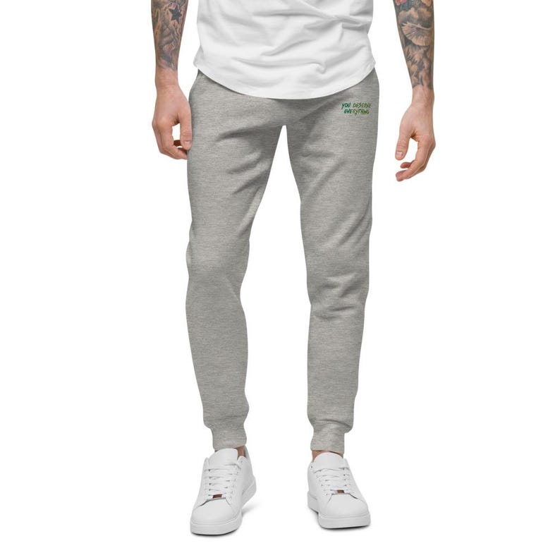 May include: Light grey sweatpants featuring a small green embroidered text on the upper right thigh that reads "you deserve everything". The sweatpants have a tapered leg design with ribbed cuffs at the ankles and a white t-shirt tucked in above.