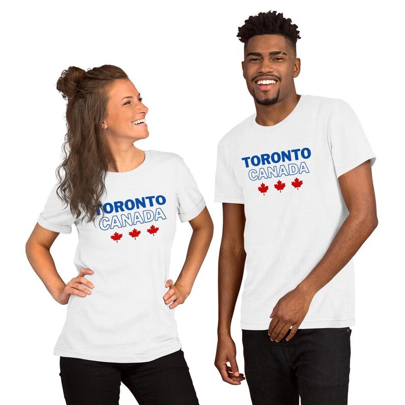 Op de afbeelding: Een wit T-shirt met korte mouwen voorzien van de blauwe tekst TORONTO en de wit omlijnde tekst CANADA, vergezeld door drie kleine rode esdoornblad-iconen die horizontaal onder de tekst op de voorzijde zijn geplaatst.