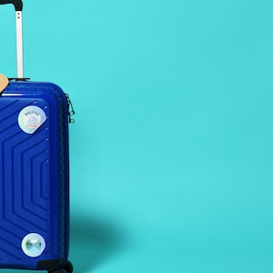 12 Canada cities as glitter pin badges - transparent PNG + printable A4 sheet. Print at home on sticker paper, cut, and use for journals, laptops, water bottles.Stickers are on a blue suitcase by Alter Standard.