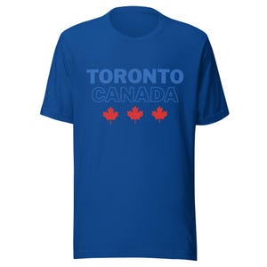 Op de afbeelding: Een koningsblauw t-shirt met korte mouwen en de tekst TORONTO CANADA in een lichtblauw lettertype. Onder de tekst staan drie rode esdoornbladpictogrammen horizontaal gerangschikt over het midden van de borst van het kledingstuk.