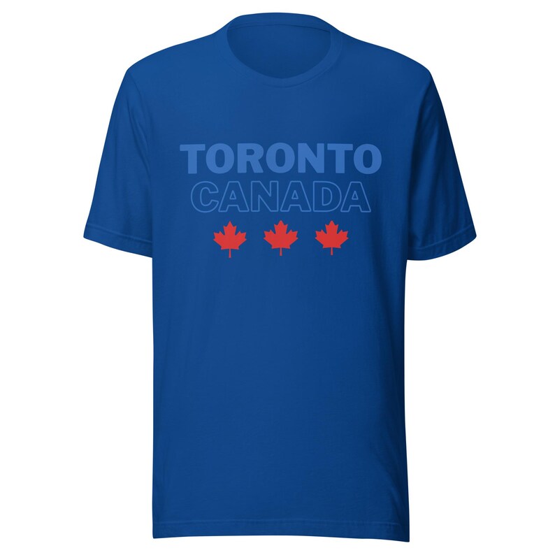 Op de afbeelding: Een koningsblauw t-shirt met korte mouwen en de tekst TORONTO CANADA in een lichtblauw lettertype. Onder de tekst staan drie rode esdoornbladpictogrammen horizontaal gerangschikt over het midden van de borst van het kledingstuk.