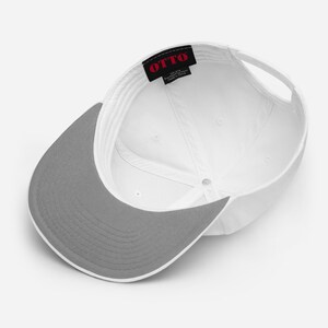 May include: A white baseball cap viewed from the inside, featuring a grey brim and a black interior label with the red text OTTO. The cap has a structured crown with white stitching and a white adjustable strap at the back for a secure fit.