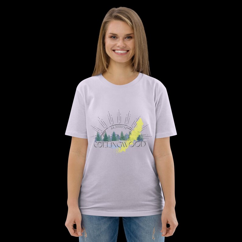 Pode incluir: Uma t-shirt de manga curta em roxo claro com uma impress&atilde;o gr&aacute;fica de pinheiros verdes, um raio amarelo e raios de sol. O texto "COLLINGWOOD" est&aacute; impresso em letras azul-escuras abaixo das &aacute;rvores com as coordenadas 44.5000&deg;N.