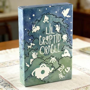 May include: A rectangular box with a watercolor design. The box features the text "Lil Cryptid Oracle" in white, surrounded by whimsical white illustrations of creatures and a bird against a blue and green background. The artist's signature is visible.