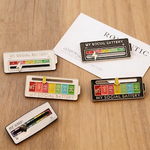 May include: Several enamel pins with the text "MY SOCIAL BATTERY" and "MY MOOD". The pins feature a sliding scale design with indicators for social energy levels. Colors include black, white, red, yellow, green, and blue.