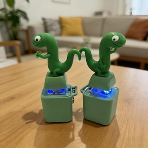 May include: Two green dinosaur-shaped desk lamps. Each lamp features a cartoon dinosaur figure atop a square base. The bases are light green and emit a blue glow. The lamps are placed on a wooden surface.