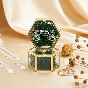 Peut inclure: Une boîte à bagues hexagonale en verre avec un cadre doré, remplie de mousse verte. La boîte est ouverte, révélant le texte "Martha Richard 12.10.2026" en blanc. Deux alliances dorées sont devant la boîte.