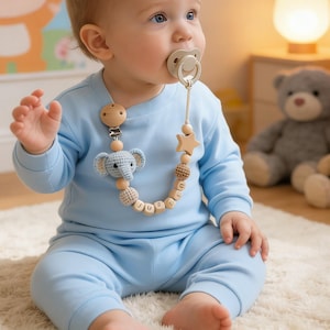 May include: A baby wearing a light blue outfit and a pacifier. The pacifier is attached to a wooden clip with a crocheted blue elephant, wooden beads, and a star. The baby is sitting on a white rug.