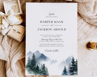 Puede incluir: Invitación de boda con los nombres Harper Mays y Jackson Arnold, y detalles del evento. La invitación tiene un diseño de montaña y bosque en acuarela. Incluye una caja de anillos, anillos y un elemento decorativo dorado.