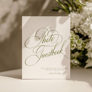 May include: A rectangular white sign with olive green cursive text that reads Photo Guestbook. Below this, smaller text reads Take a photo, place it in the book and leave a message for Abby & Garry. The sign features a smooth, matte finish.