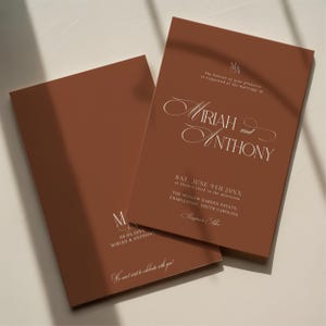 May include: Two terracotta-colored wedding invitations with elegant white script. The text includes the names Mariah and Anthony, the date, time, and location of the ceremony. The invitations are rectangular and feature a minimalist design.