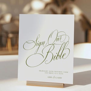 May include: A white rectangular sign with olive green cursive text that reads Sign Our Bible. Below this, smaller text reads Highlight your favourite verse and write us a note, followed by the names Abby & Garry in a simple sans-serif font.