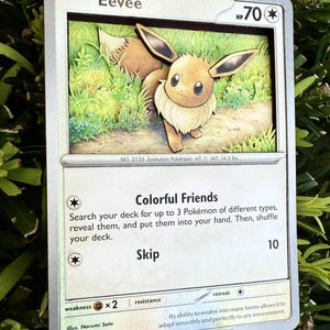 May include: A framed Eevee Pokémon card. The card features a 3D rendering of the Eevee character in a grassy setting. The card text includes "Eevee", "Colorful Friends", and "Skip". The card measures 1 foot in height and weighs 14.3 pounds.