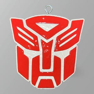 May include: A red and white Transformers logo ornament with a silver metal loop for hanging. The emblem is a stylized shield shape with a glossy finish, perfect for fans of the franchise.