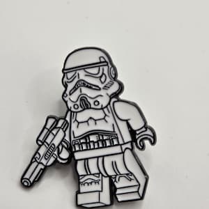 May include: A white and black enamel pin featuring a Stormtrooper character holding a blaster. The pin is in a block figure style, with detailed outlines and a glossy finish. The design is based on the iconic Star Wars character.
