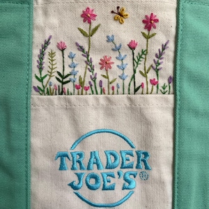 Może przedstawiać: Zbliżenie płóciennej torby z białą kieszenią z haftowanymi kwiatami i motylem. Torba ma miętowy zielony brzeg, a słowa "TRADER JOE'S" są wyhaftowane na miętowo-niebiesko.
