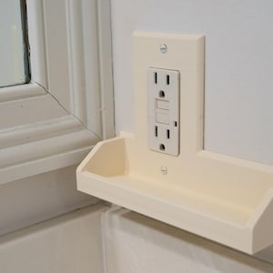 May include: Off-white electrical outlet cover with a built-in shelf. The outlet has two standard receptacles and a reset button. The shelf is rectangular with a slightly curved front edge. The cover is mounted on a white wall.
