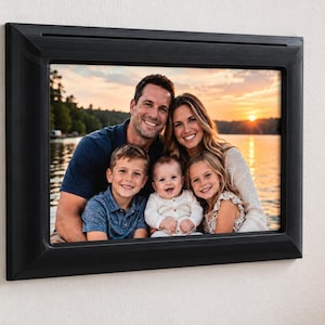 May include: A rectangular digital photo frame with a black plastic border mounted on a wall. The screen displays a color photograph of a family of five, including two adults, a young boy, a baby, and a young girl, posing outdoors at sunset.
