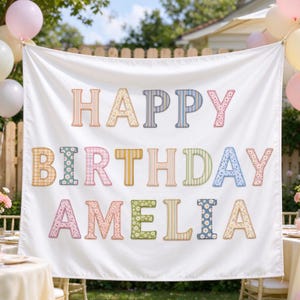 May include: A white fabric banner featuring the text HAPPY BIRTHDAY AMELIA in large, multi-colored, patterned fabric letters. Each letter displays a unique print, including stripes, gingham, and floral designs in pastel and muted tones.