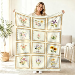 Pode incluir: Cobertor creme com uma grade de desenhos florais em molduras de selos postais. Cada moldura mostra um arranjo floral em aquarela, com nomes como "Nora" e "Ruby" abaixo. O cobertor é segurado por uma pessoa.